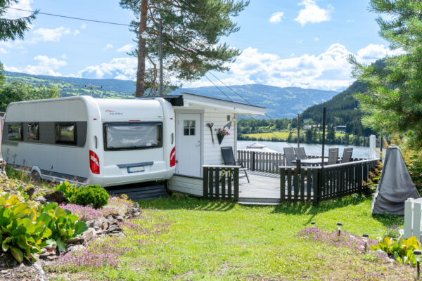 Campingvogn med spikertelt og inngjerdet terrasse på campingplass. Omringet av blomsterbedd og vakker natur. Foto
